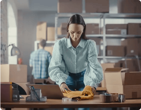 Young lady folding a yellow sweater next to a laptop in a warehouse